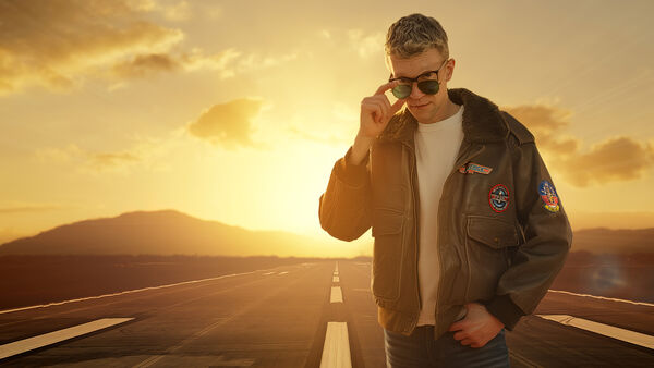 Performer Richard Marsh stands on an airport runway at sunset, wearing a brown flight jacket with military patches and peering over a pair of aviator sunglasses.